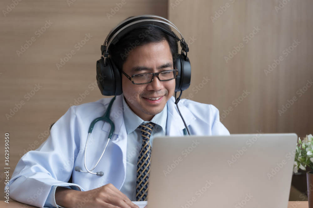 亚洲男医生戴着耳机在办公桌工作，对着镜头微笑，视频通话咨询