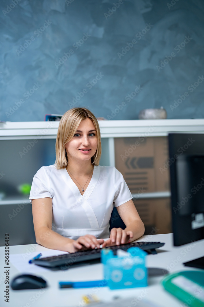 在医务室里，穿着磨砂服的女护士在电脑前工作。看着镜头。快乐的女人