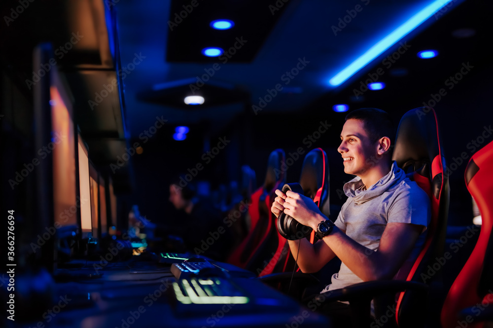 微笑的少年在网吧里戴着耳机看着电脑屏幕，肖像。