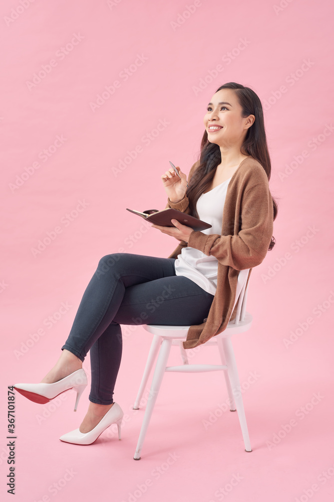 微笑的可爱女孩坐在粉色墙上的椅子上写日记