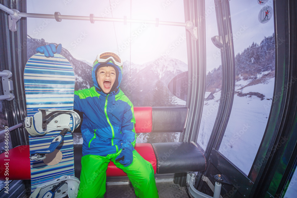 Happy boy with snowboard screaming from happiness sitting in the ski lift cabin on winter resort