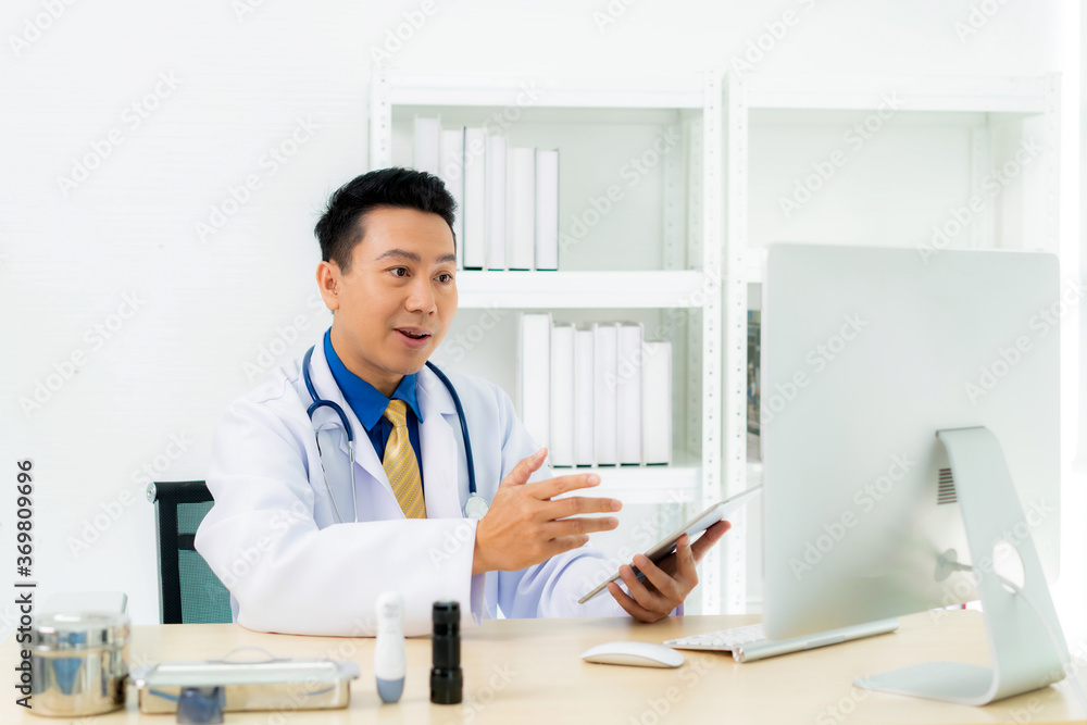 Asian man doctor wears white coat and headset speaking videoconferencing on laptop computer using on
