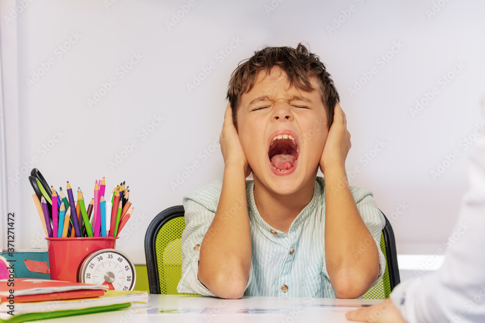 自闭症男孩坐在cl的桌子旁，在ABA发育治疗过程中痛苦地尖叫并闭上耳朵