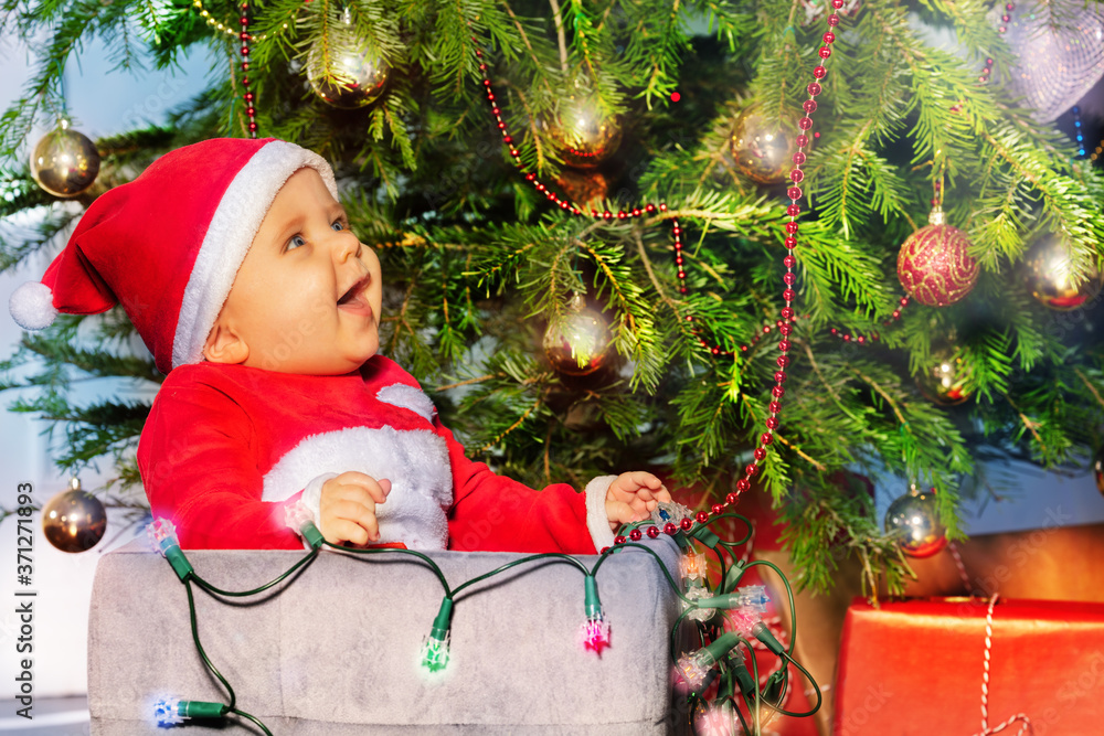 戴着圣诞老人帽子、拿着发光的灯坐在圣诞树下的盒子里大笑的男婴