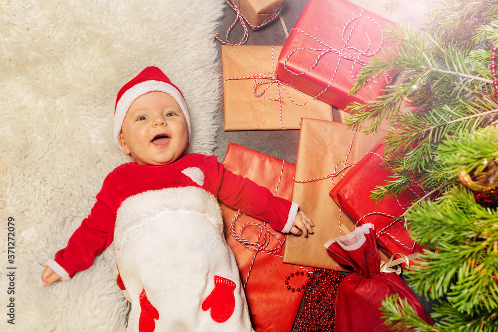漂亮的男婴躺在地板上，放在一堆礼物盒子里，微笑着抬头看着新年树