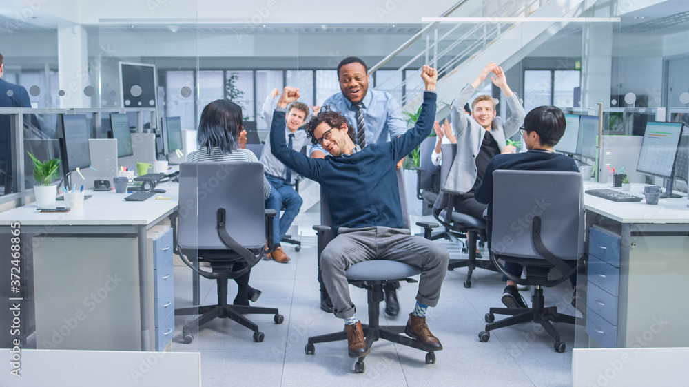 Cheerful Employee Pushes His Handsome Colleague on a Chair Between Rows of Desks with Diverse Busine
