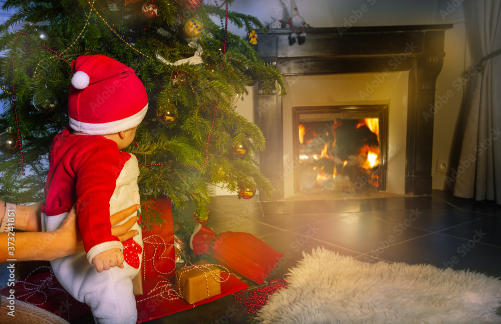 可爱的蹒跚学步的男婴在圣诞树前看着母亲手中的壁炉