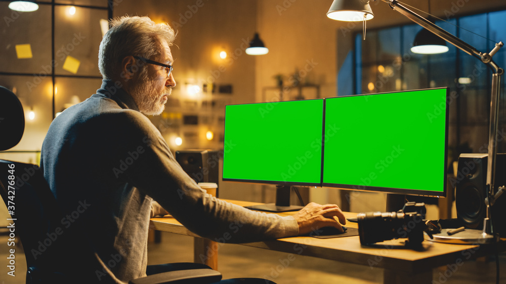 Creative Middle Aged Designer Sitting at His Desk Uses Desktop Computer with Two Green Mock-up Scree