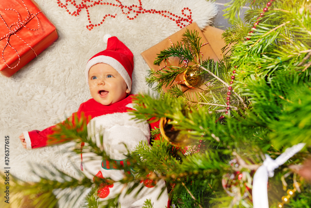 快乐的蹒跚学步的小男孩躺在地毯上，张着嘴笑着看着装饰过的圣诞节