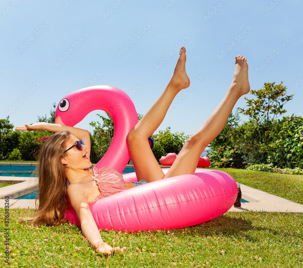戴着太阳镜的女孩在游泳池附近的粉红色火烈鸟上摆出有趣的摔倒姿势