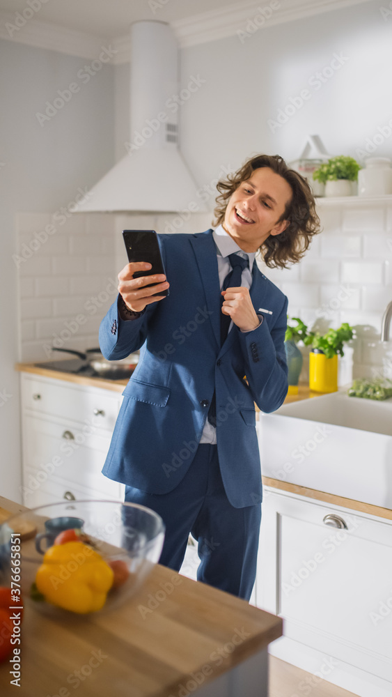 快乐的长发年轻人穿着蓝色西装在厨房里用智能手机。他是