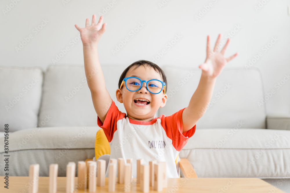 快乐、聪明、书呆子气十足的蹒跚学步的亚洲男孩在家玩木制玩具积木获胜时举起了手。男孩chi