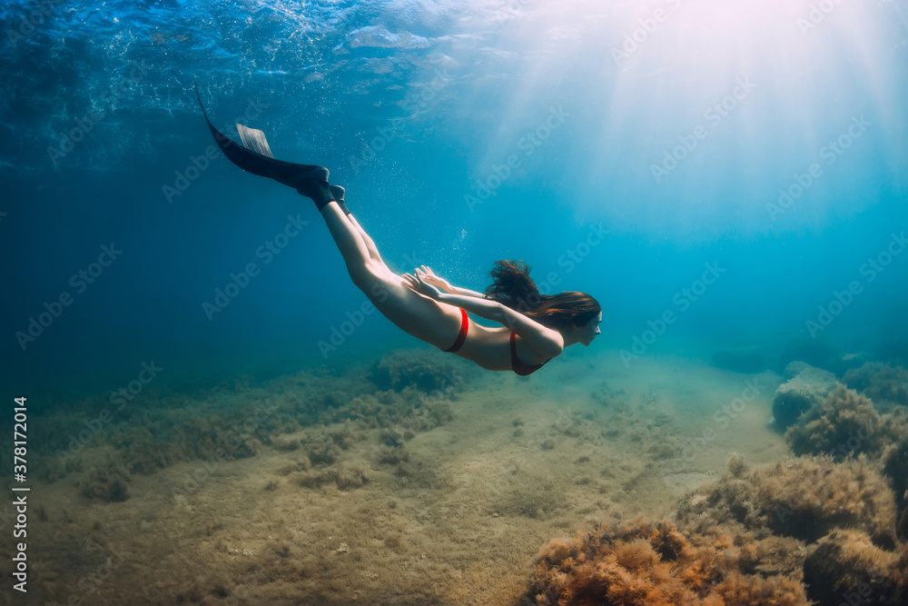 Freediver woman in bikini glides in blue sea and sun rays. Freediving with fins underwater in ocean
