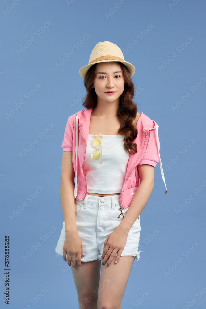 Portrait of young Asian woman wearing hat, t-shirt, short jeans and pink jacket isolated over blue b
