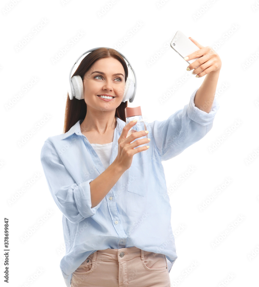 Young woman with bottle of water taking selfie on white background