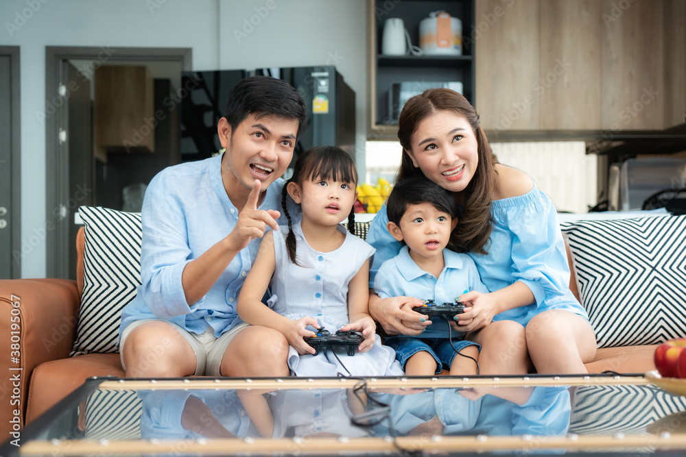 Happy asian family with father, mother, daughter and son playing games using joystick having funny m