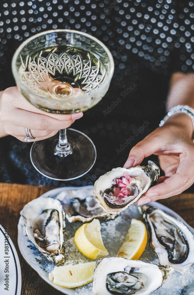 Hands of woman holding glass of champagne and fresh oyster with lemon juice in fish restaurant, sele
