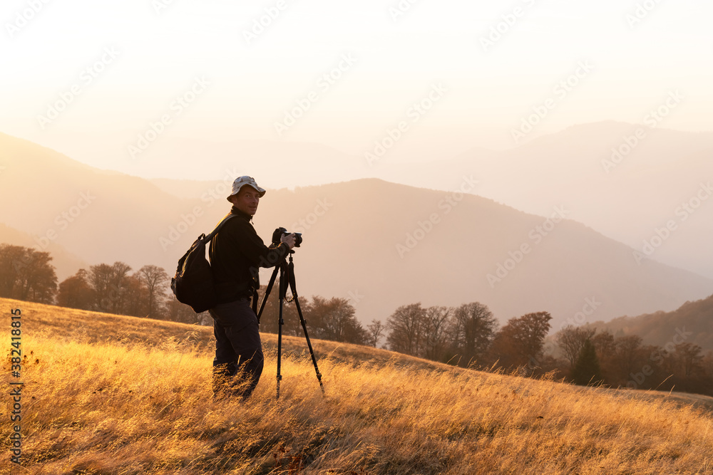 Photographer taking photo with tripod of autumn landscape with foggy peaks and orange trees. Ukraini