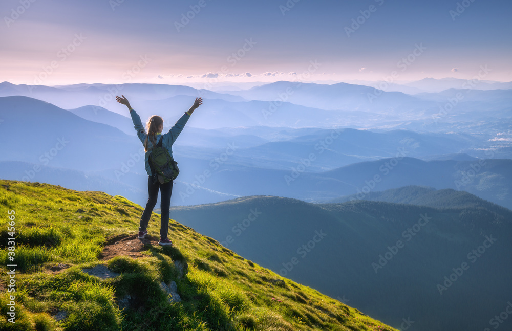 Happy woman with backpack and raised up arms on the peak and beautiful mountains in fog at sunset. A
