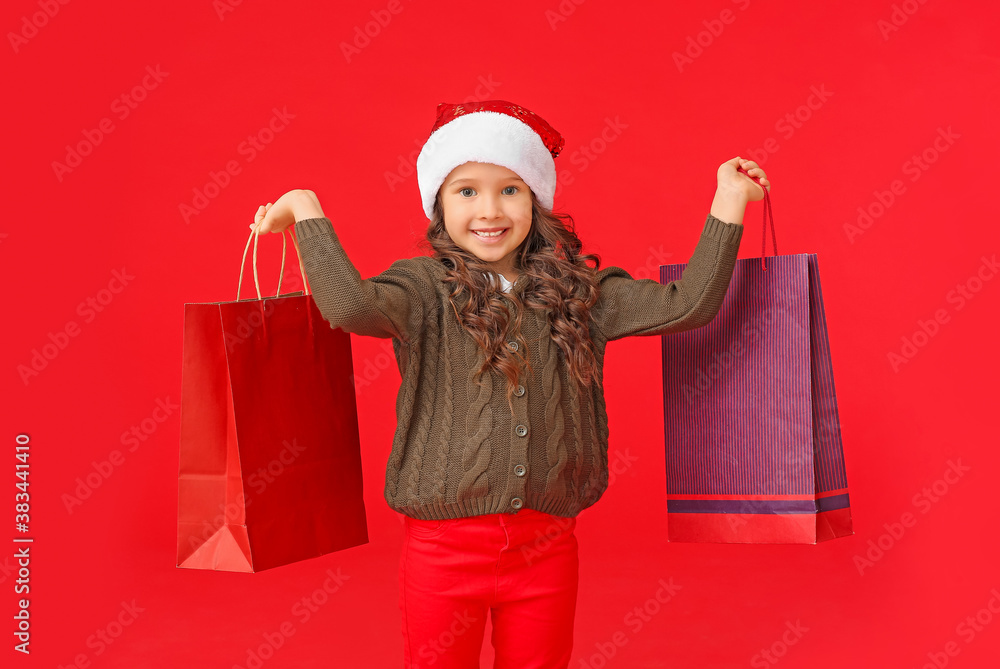 Cute little girl in Santa hat and with shopping bags on color background