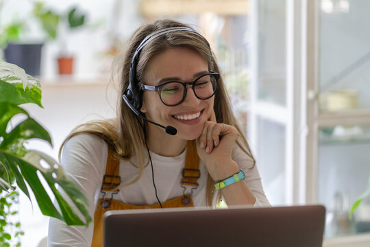 Smiling gardener millennial woman wear headphones talk video calling, using laptop, communicating wi