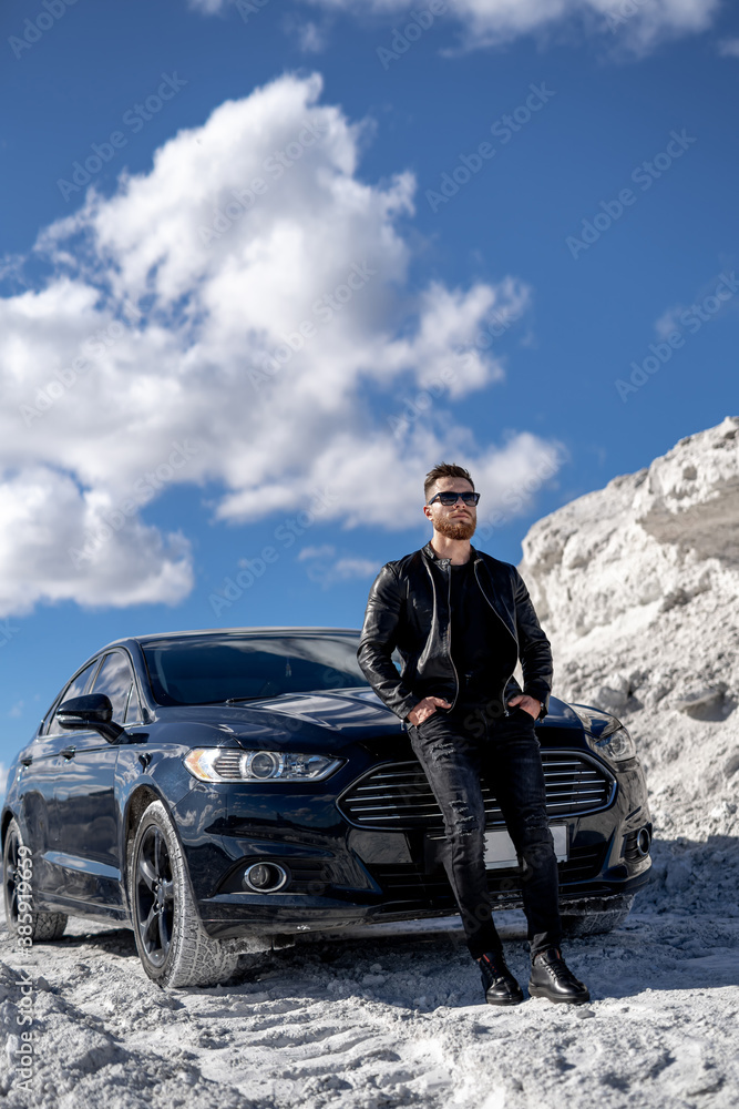 Portrait of a stylish bearded man in trendy sunglasses and leather jacket stands near black car. Fas