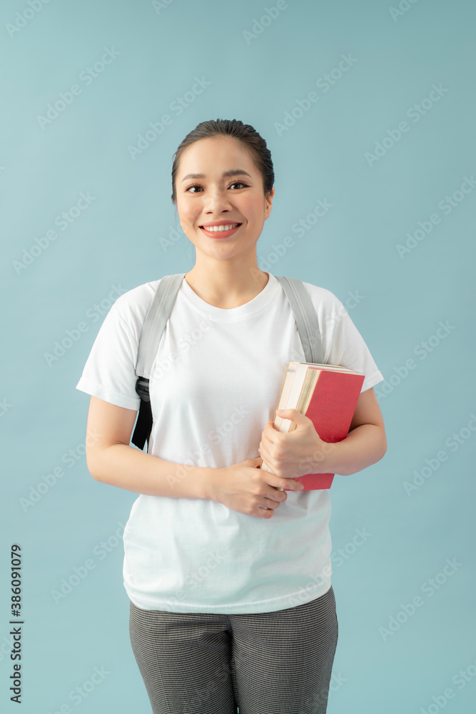 Portrait of a smiling asian student girl wearing backpack and holding books isolated over blue backg