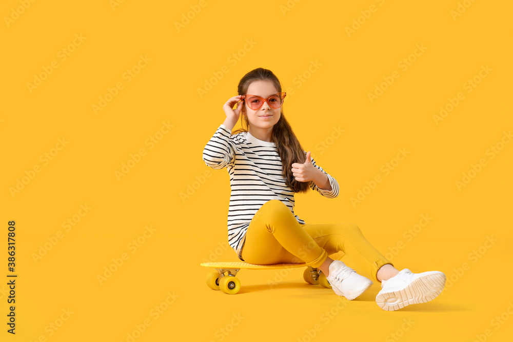 Cute little girl with skateboard showing thumb-up gesture on color background