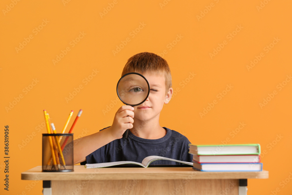 Cute pupil sitting at desk against color background