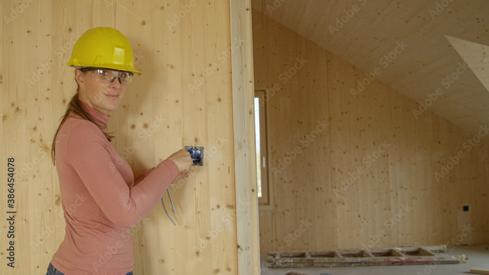 PORTRAIT: Young female electrician screws an outlet frame onto a wooden wall