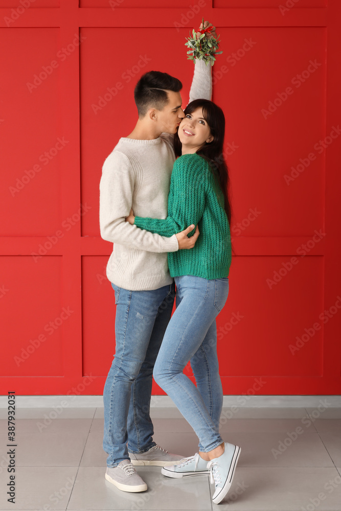 Young man kissing his wife under mistletoe branch near color wall