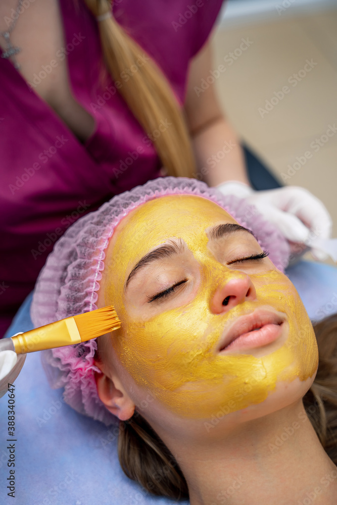 Cosmetologist applies golden mask with brush on face of woman. Facial skin and anti-aging treatment.
