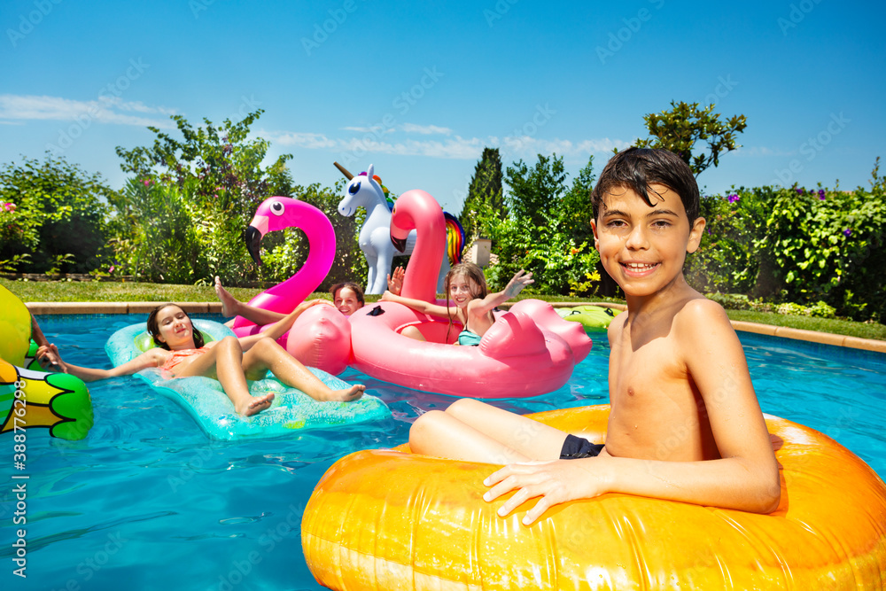 Smiling boy in group of children play, have fun in swimming pool outside pose with inflatable doughn