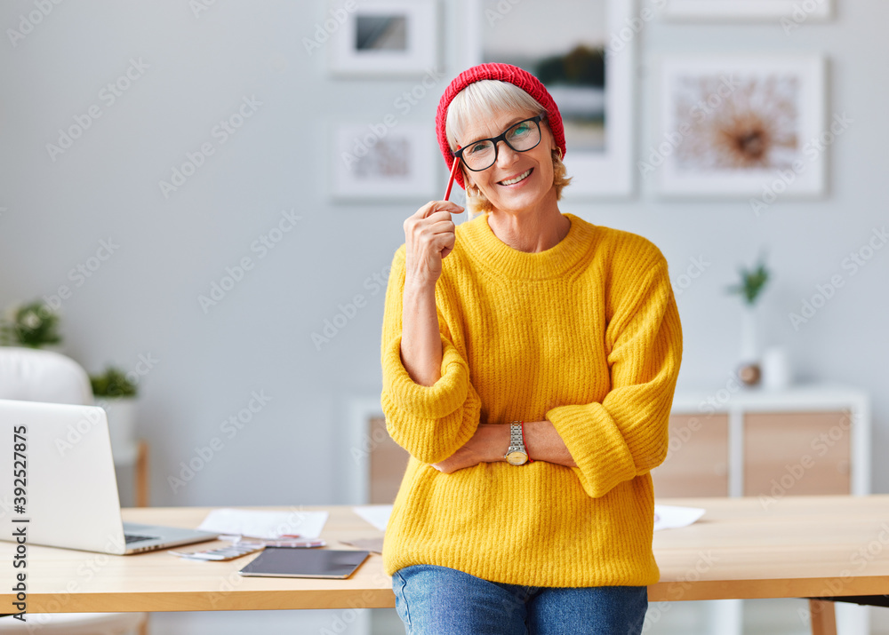 戴着红帽子的快乐老年女性创意设计师拿着笔记本电脑在工作场所微笑，ske