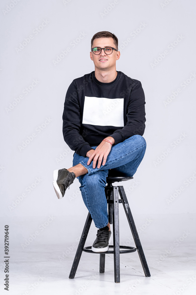 Smiling guy sits on high chair with leg on knee. White background and casual clothes. One head on kn