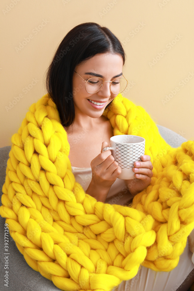 美丽的年轻女子，穿着温暖的格子布，坐在彩色背景的扶手椅上喝茶