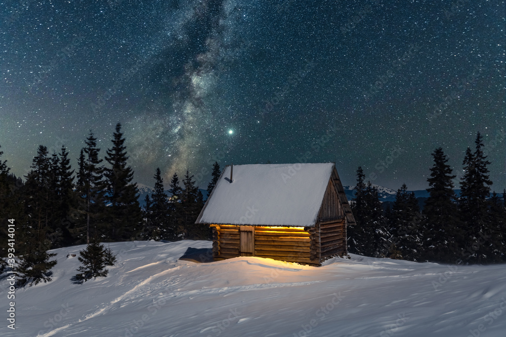 Fantastic winter landscape with wooden house in snowy mountains. Starry sky with Milky Way and snow 