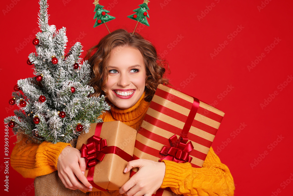 快乐的女人微笑着，手里拿着一棵小圣诞树和红色背景上的礼物