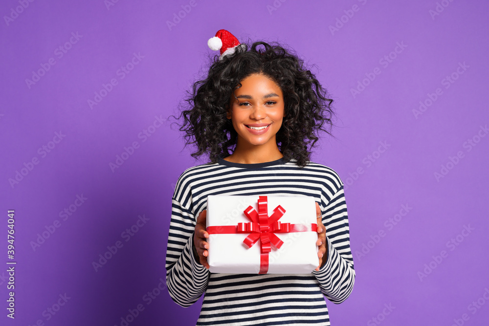 戴着圣诞老人帽、面带微笑的少数民族妇女在五颜六色的紫色背景上拿着圣诞礼物