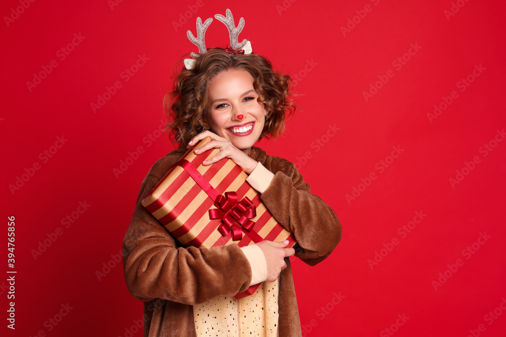 穿着圣诞驯鹿服装的可爱有趣的年轻女子微笑着，拿着五颜六色的红色礼物欢欣鼓舞