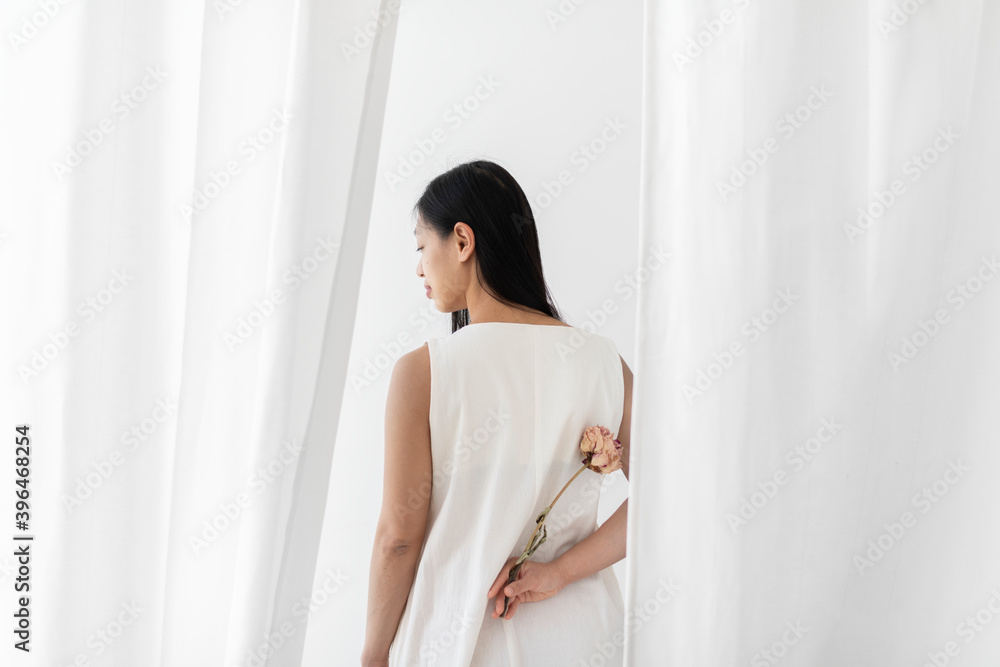 白色窗帘中，手里拿着一朵干粉色牡丹花的亚洲女人