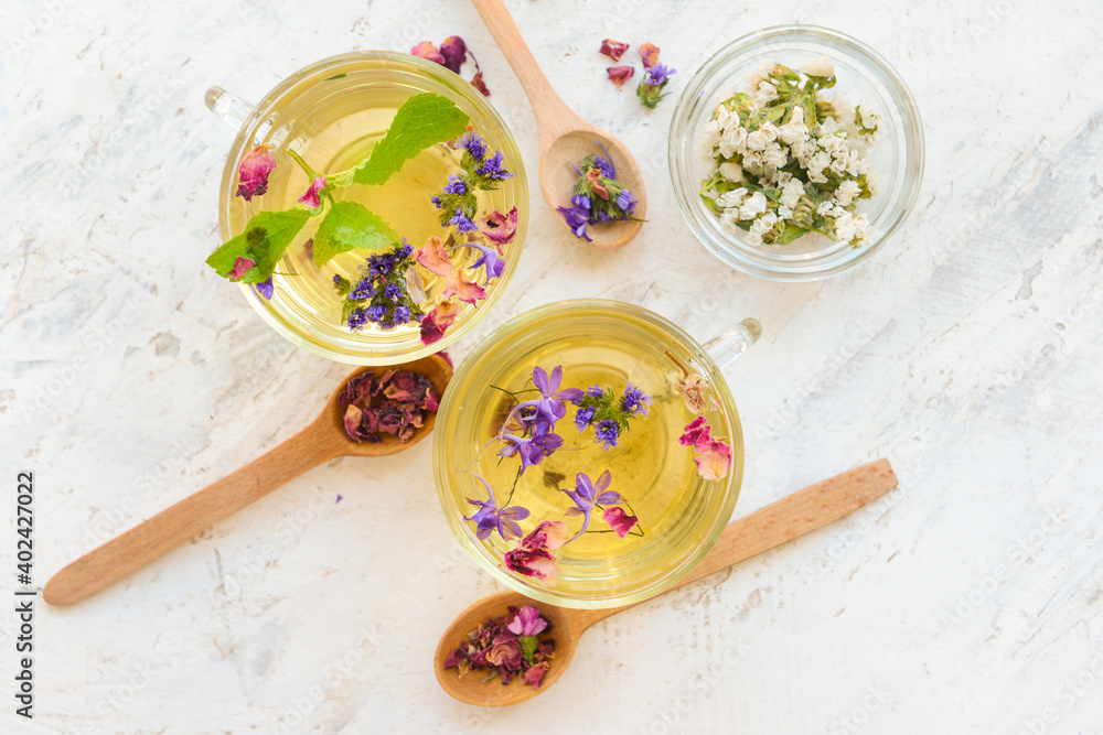 Cups with floral tea and herbs in spoons on light background