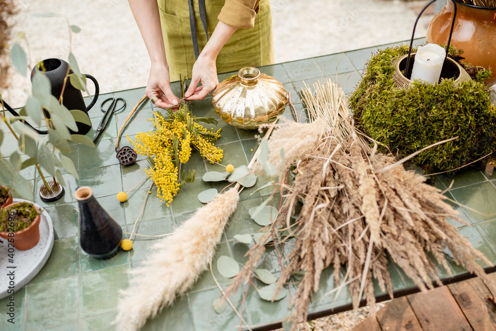 Female making compositions of dried and fresh flowers and herbs on the green table outdoors. Florist