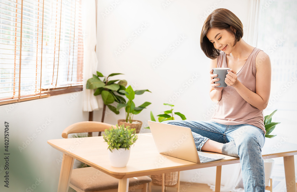 年轻美丽的亚洲女人的肖像，放松地坐在桌子上，使用电脑笔记本电脑。微笑快乐的亚洲女孩