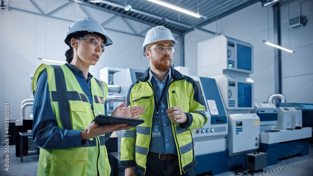 现代工厂：女项目经理和男工程师站着，穿着安全夹克，戴着安全帽，