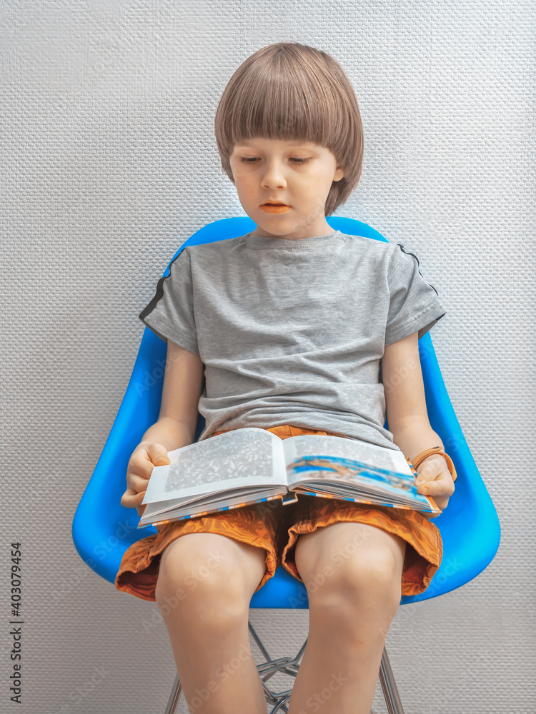 一个穿着休闲服的男孩坐在蓝色椅子上，在白墙前看书。