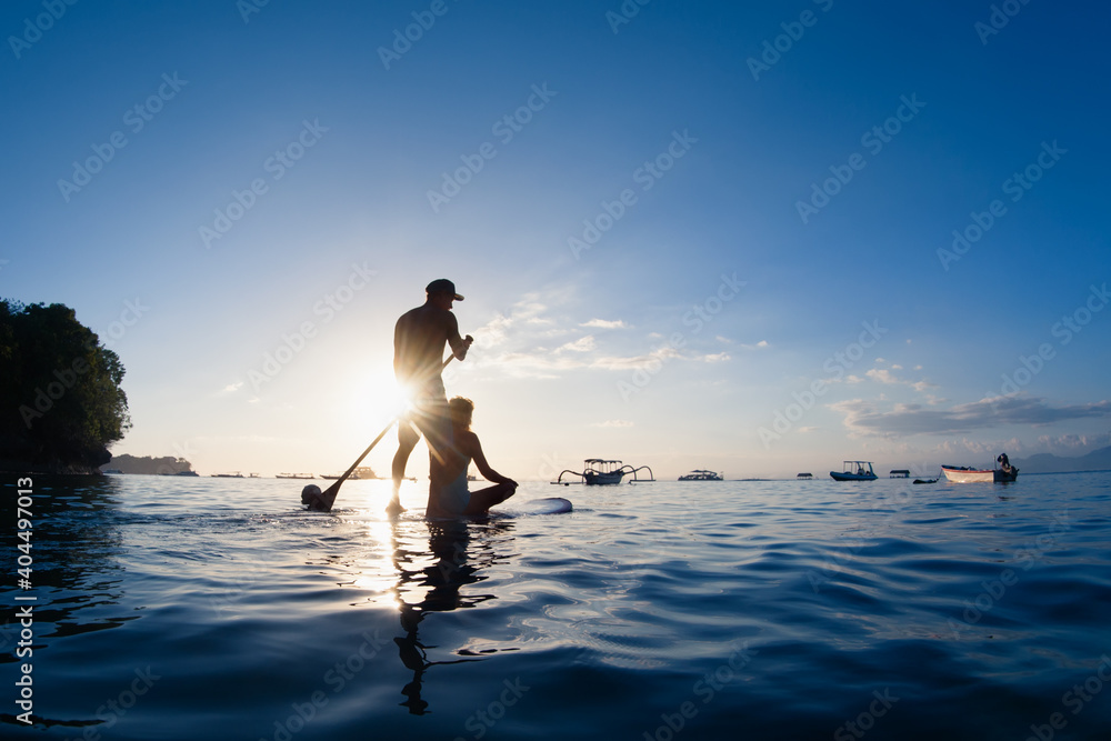 年轻幸福的夫妇在立式桨板上玩得很开心。活跃的桨板手在日落的海边划水。H