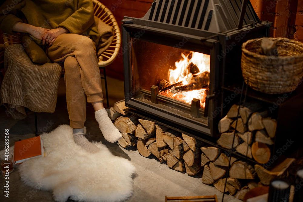 年轻的女人坐在燃烧的壁炉旁，在舒适的阁楼风格的室内享受热饮放松