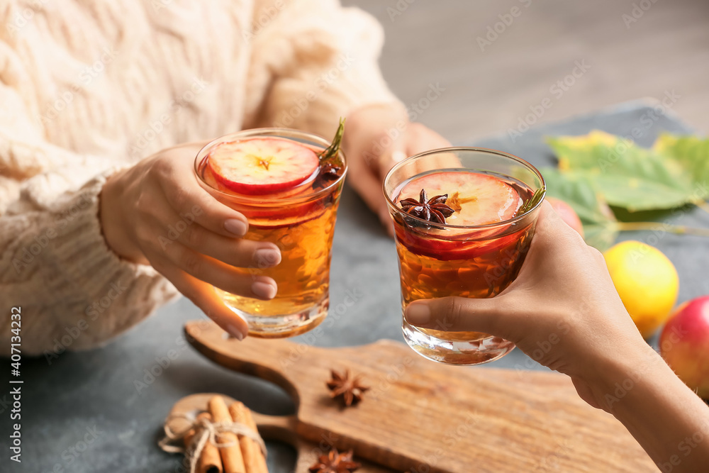 Women holding tasty drinks with spices and apple slices in glasses on dark background