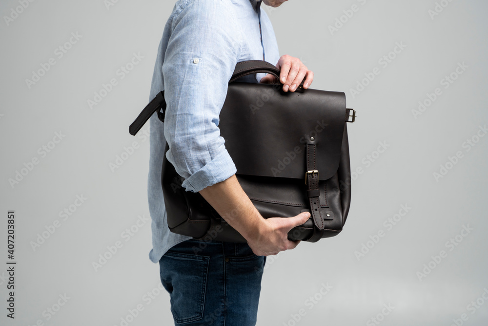 穿着蓝色衬衫和牛仔裤的男人拿着文件和笔记本电脑的棕色男士双肩皮包